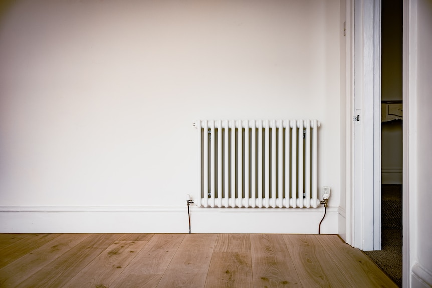 A built in central heating radiator hangs in the bottom right corner of a white wall with wooden floors beneath it