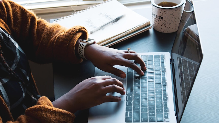 Woman typing on a laptop with a notepad and cup beside her