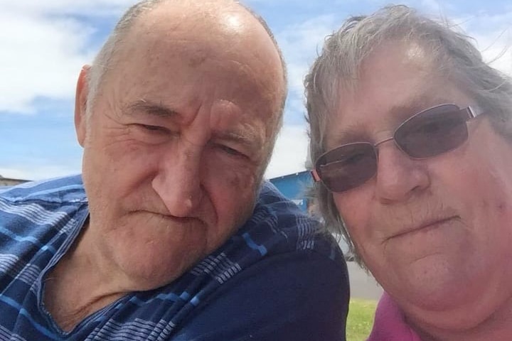 A man and woman sitting on a bench pose for a selfie, big blue sky behind them.