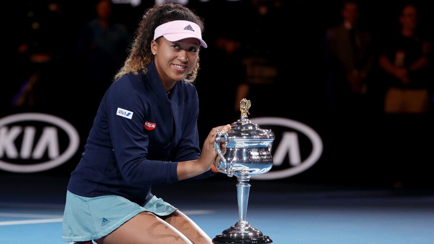 Naomi Osaka kneels on the court next to the Australian Open winner's trophy.