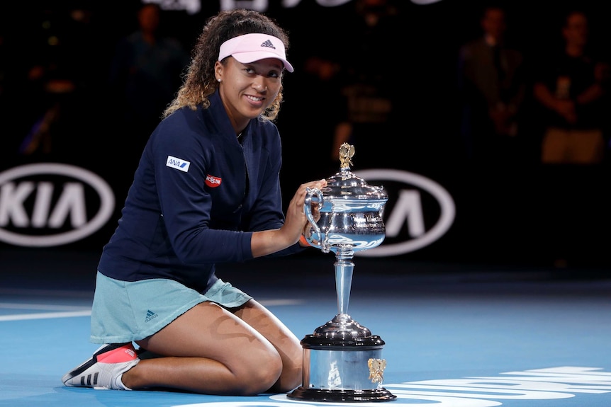 Naomi Osaka kneels on the court next to the Australian Open winner's trophy.