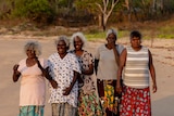 Five Burarra women in shirts and long skirts stand together on a beach in the afternoon sun.