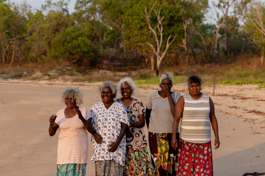 Five Burarra women in shirts and long skirts stand together on a beach in the afternoon sun.