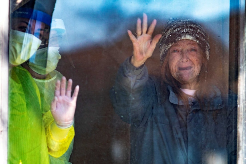 An elderly woman in a beanie waves through a window next to two people in full PPE