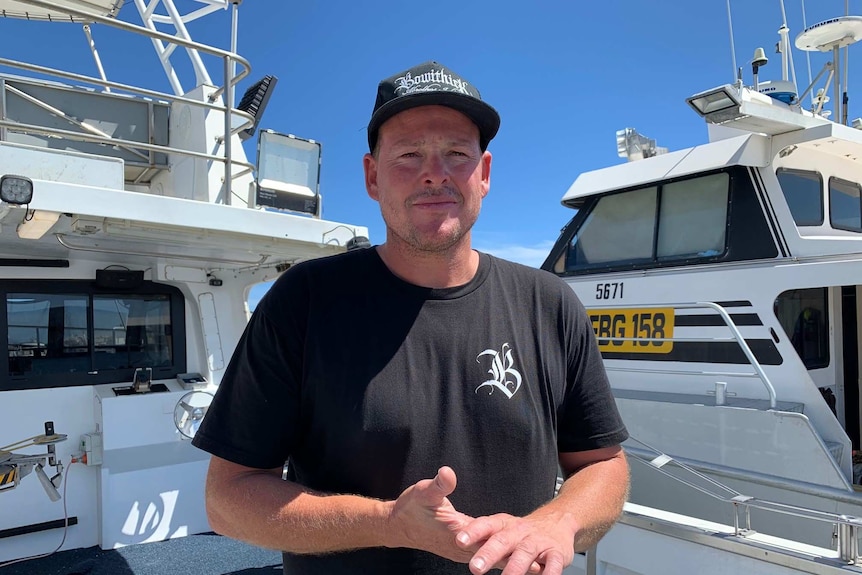 A middle-aged man stands in front of two fishing boats, squinting at the camera.