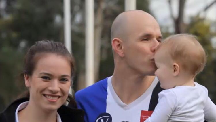 A man in a football jumper hugs a woman while holding and kissing a baby