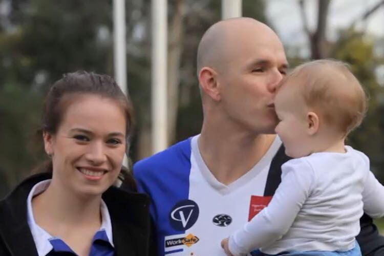 A man in a football jumper hugs a woman while holding and kissing a baby