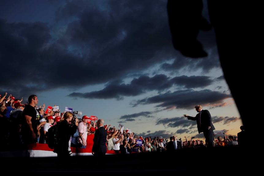 A man in a suit stands before a crowd under a darkening sky