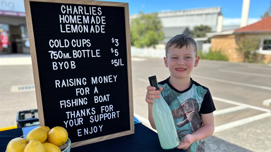 A boy stands next to a sign listing prices for the homemade lemonade he's selling at this stand. 