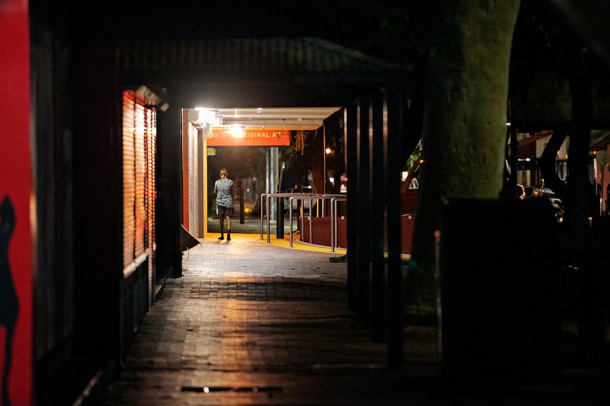 A teenager walks down a dark sidewalk with their back to the camera. 