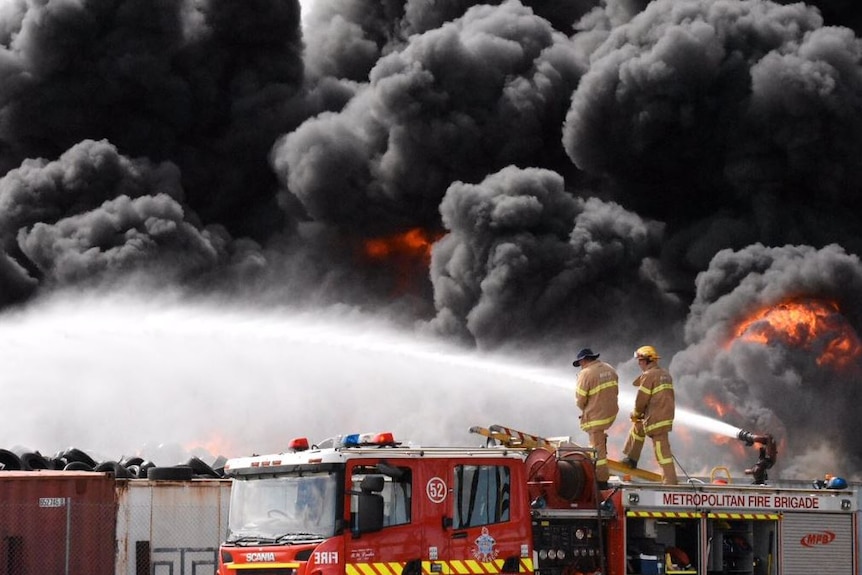 Fire crews stand on trucks near large plumes on smoke.