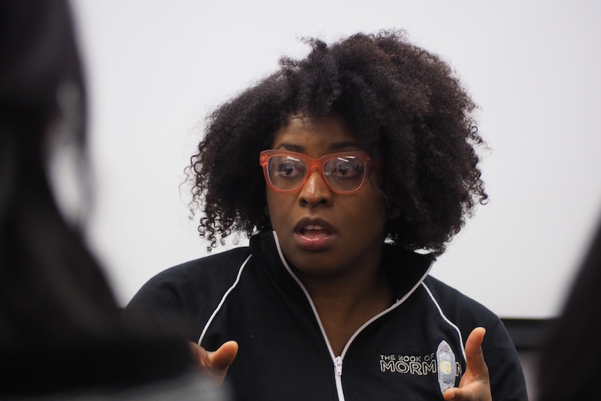 Cessalee Smith-Stovall, who is wearing big orange glasses, mid-conversation with other actors. 