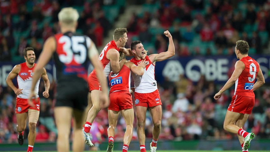 Teammates surround an AFL player who punches the air having kicked a goal.