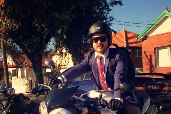 Matthew Anthony Ridley sitting on a motorcycle wearing a suit and an open-face helmet.