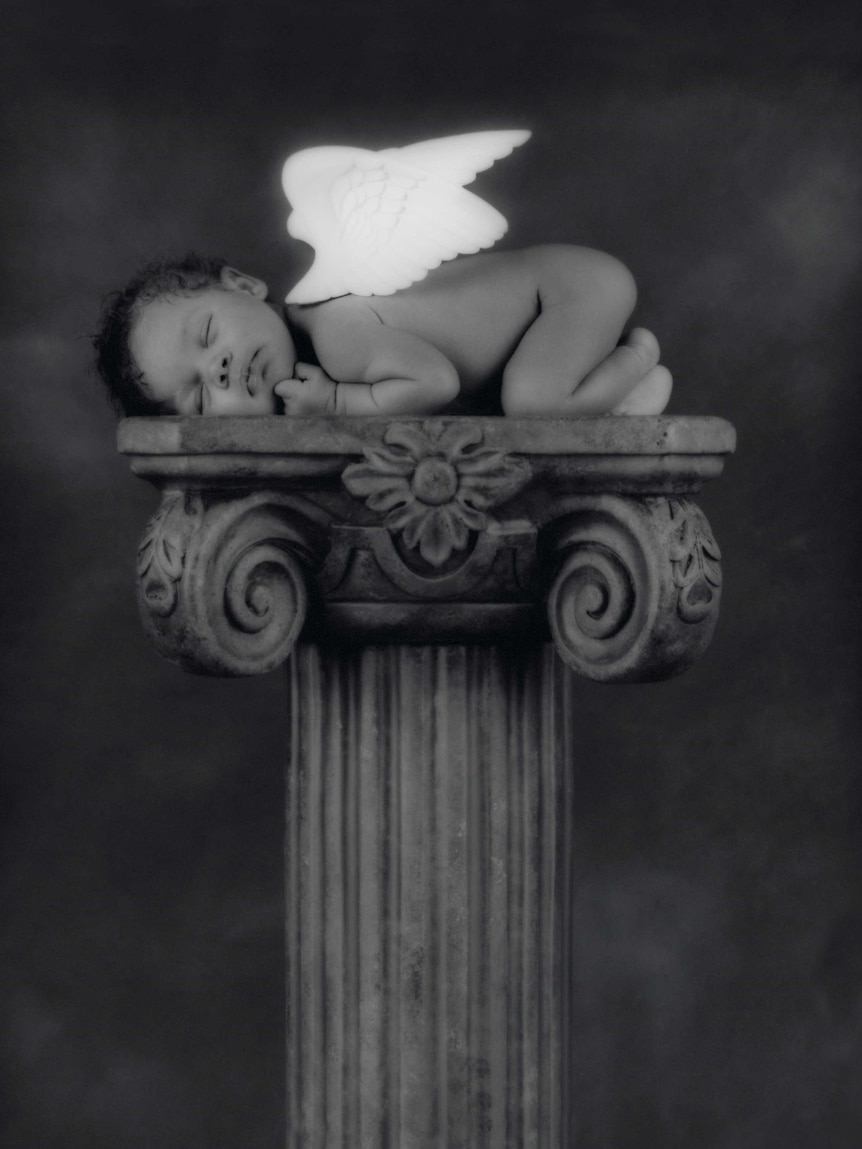A black and white photo of a baby with angel wings on a greco column/plinth by Anne Geddes