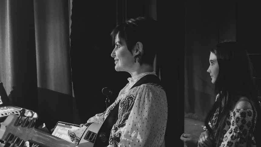 black and white photo of a woman who stands in front of a theatre curtain holding a guitar while a teenage girl stands behind