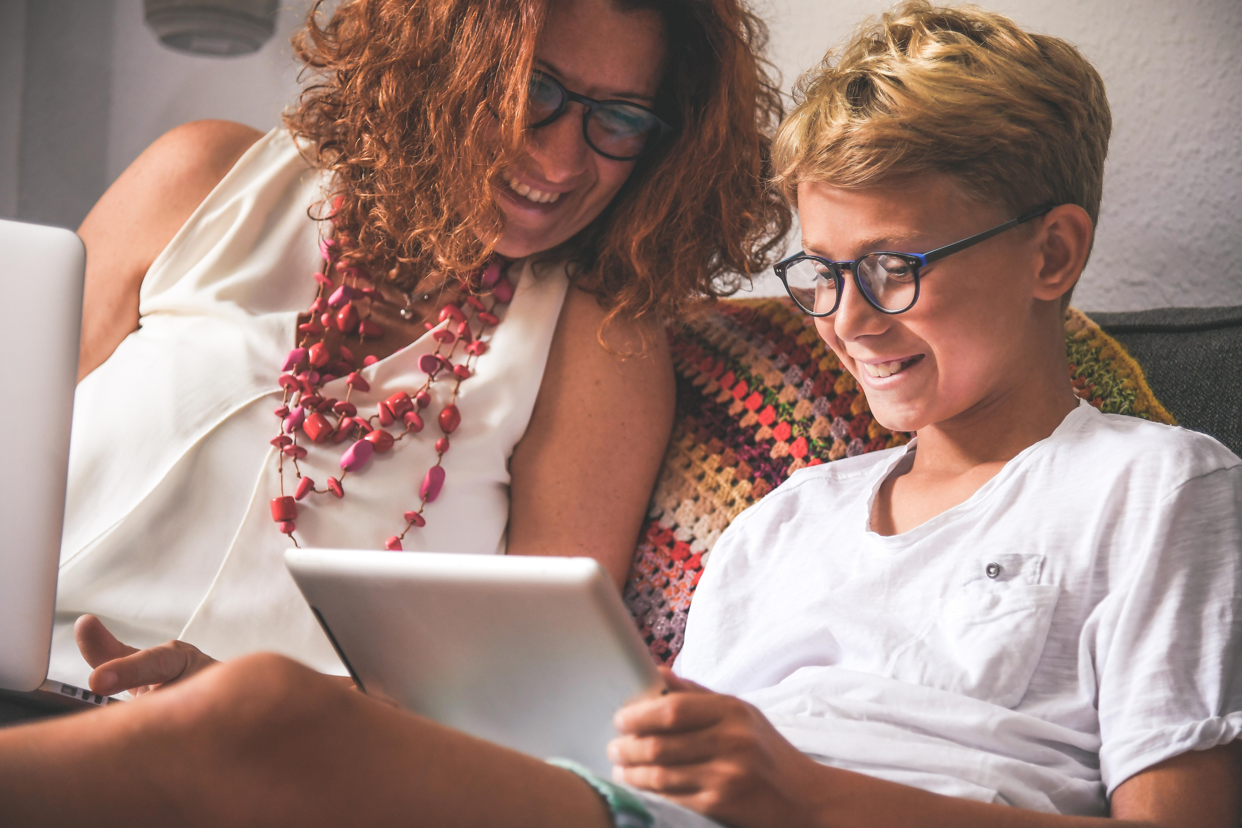 A smiling mother with a laptop and her son with a tablet. They sit on a couch and the mother looks over to the son’s tablet.
