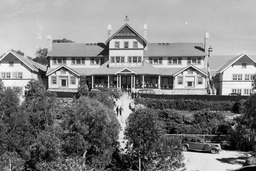 A black and white picture of a multi-story ski chalet surrounded by native trees and manicured hedges 