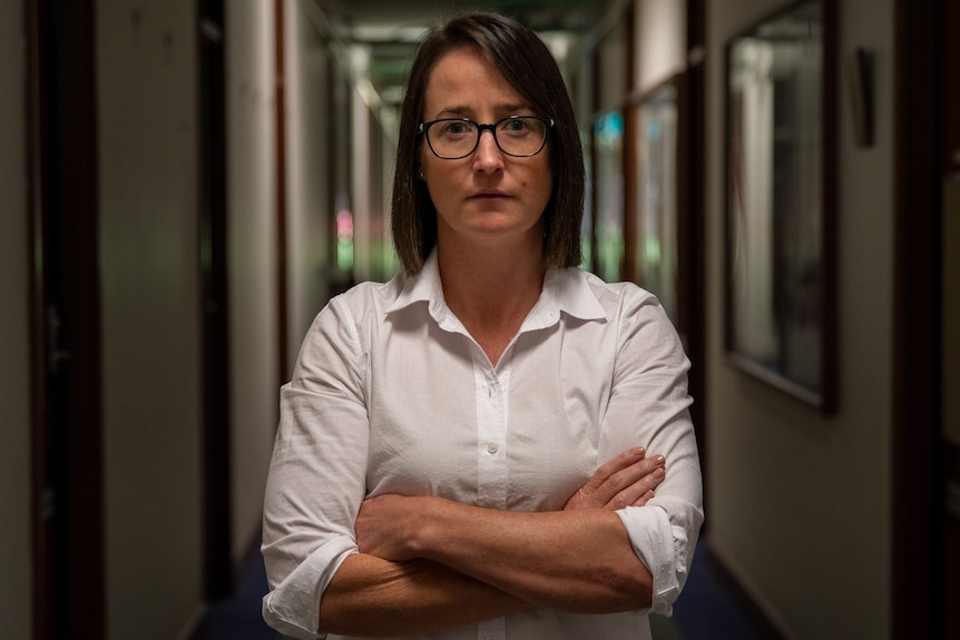 Woman with dark hair and glasses looking to camera standing in dimly lit corridor. 