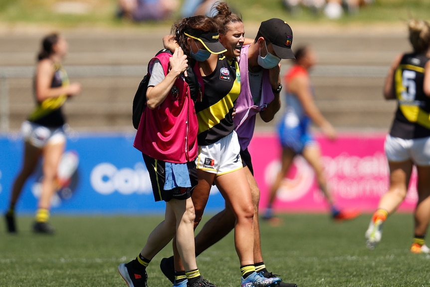 An injured Richmond AFLW player is helped off the field by two of the club's medical staff.