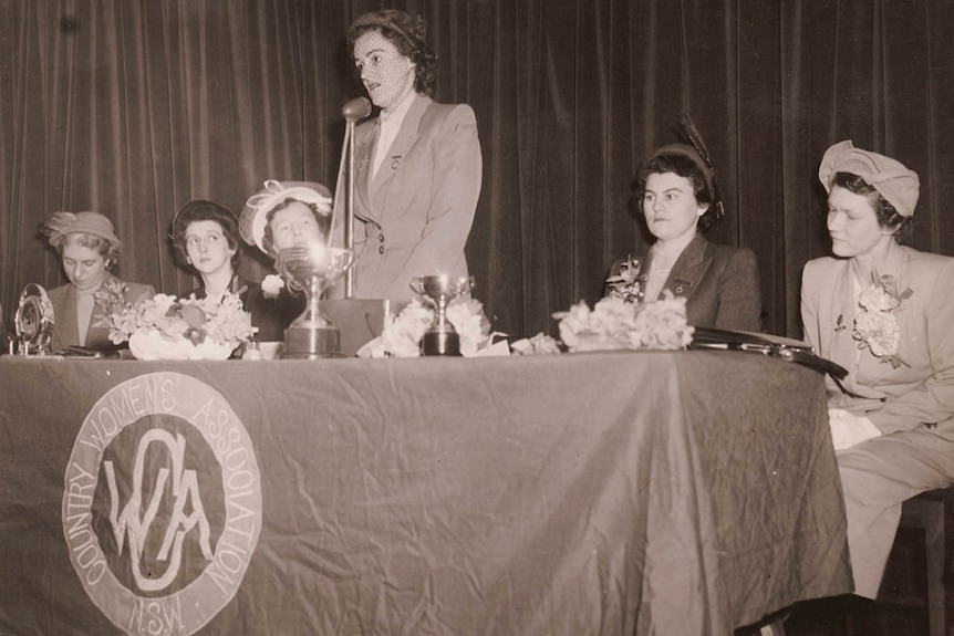 Black and white picture of well dressed women standing behind a table for the CWA
