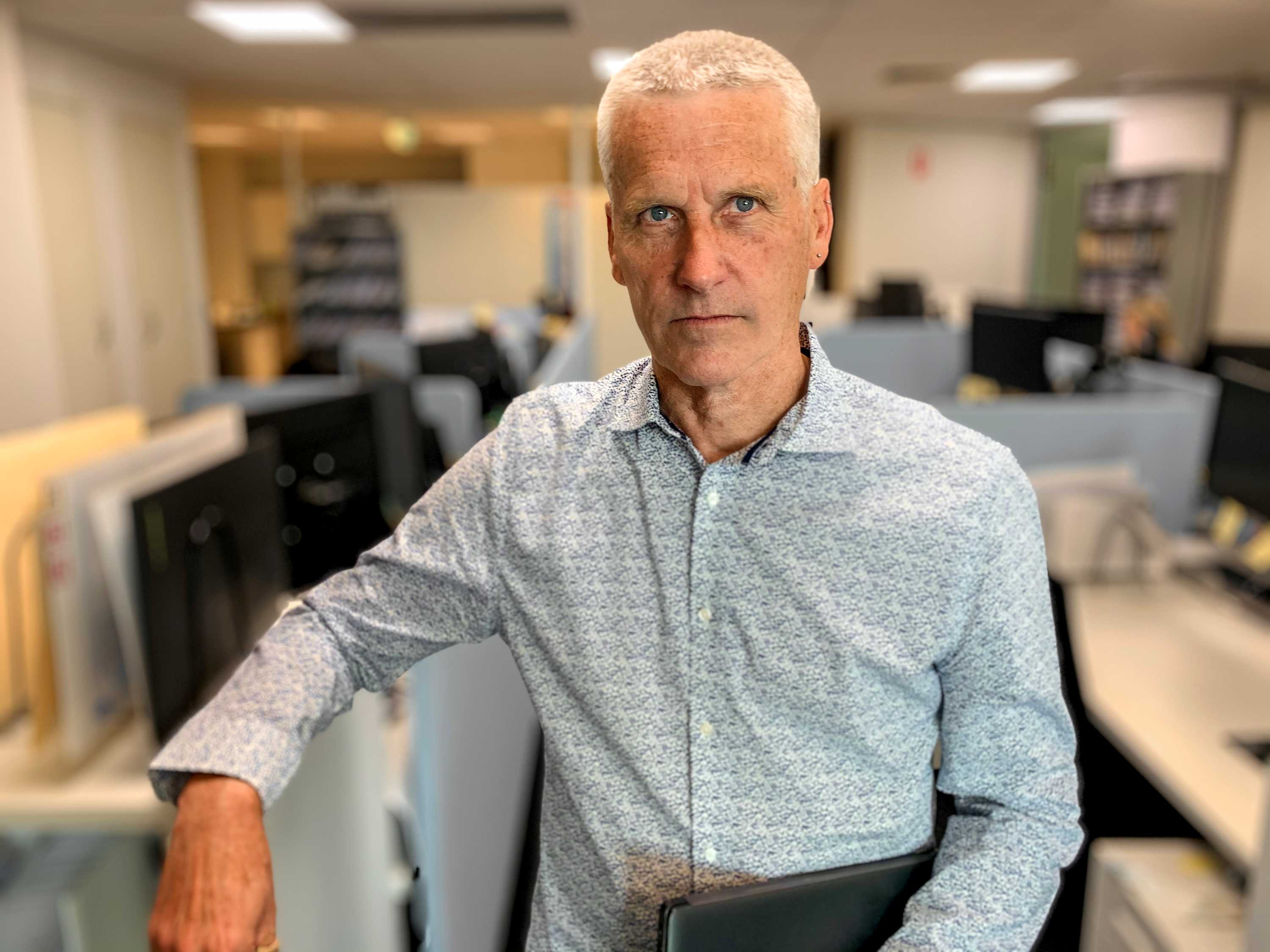 Man with white hair wearing light blue shirt stands in office with desks visible behind him