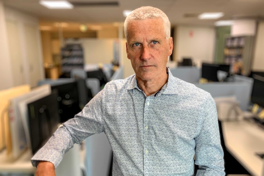 Man with white hair wearing light blue shirt stands in office with desks visible behind him