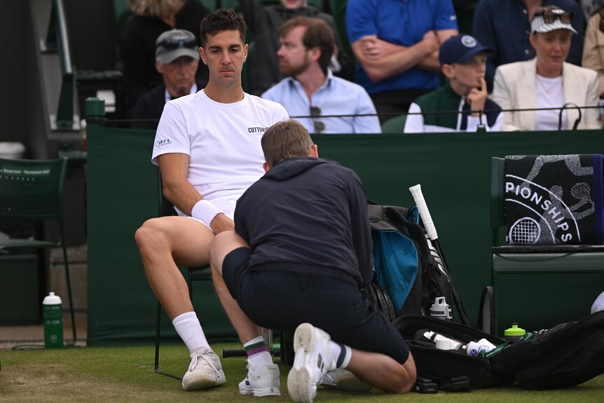 Australia's Thanasi Kokkinakis sits in his chair at Wimbledon looking sad as a trainer examines his injured knee.