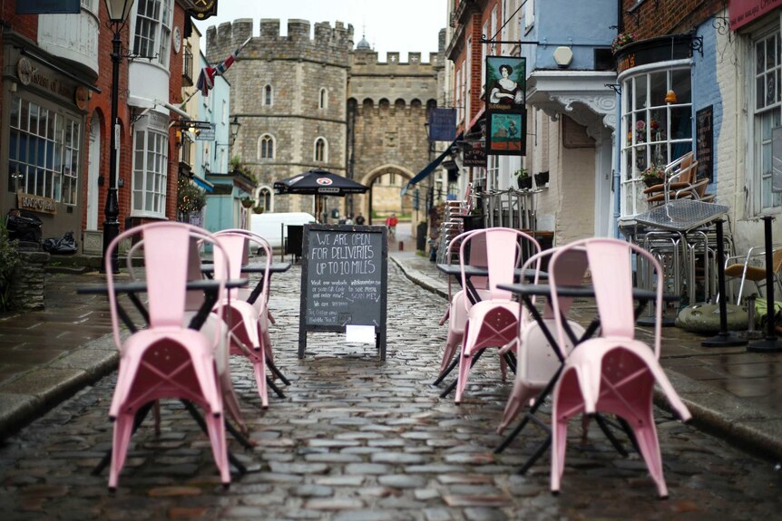 A cobble-stoned street with a sign and pink chairs titled in front of a venue, with castles and English facades in background.