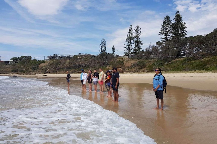 Tourists stand at water's edge on Coffs Head beach with Unkya Cultural Eco Tour guide.