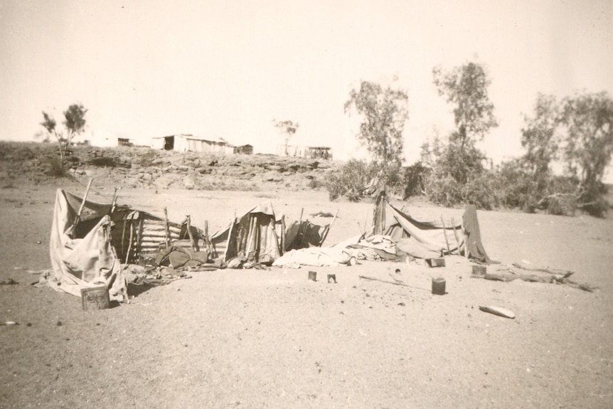 A black and white photo showing corrugated iron and hessian strung up in a creek bed, a larger tin structure on the ridge behind