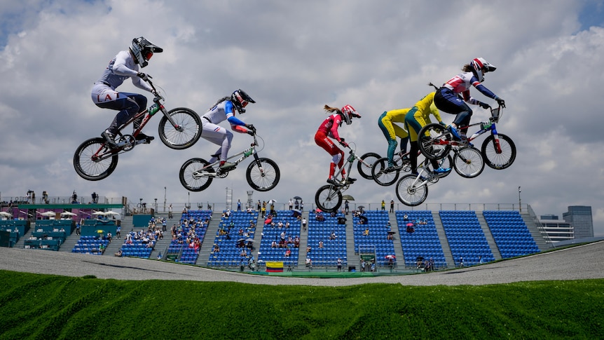 Six riders are high in the air as they go over a big jump in the BMX racing, with a stand in the background.