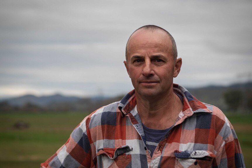 Craig McKimmie has stares at the camera, wearing a red, blue and white flannelette shirt, green paddocks and hills behind.