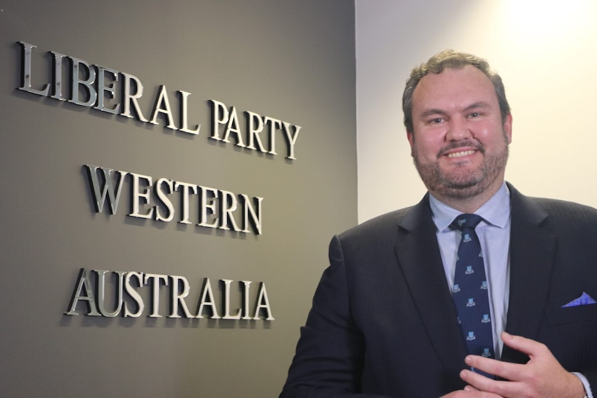 Richard Wilson en costume-cravate debout devant des pancartes qui disent "Parti libéral d'Australie occidentale" 