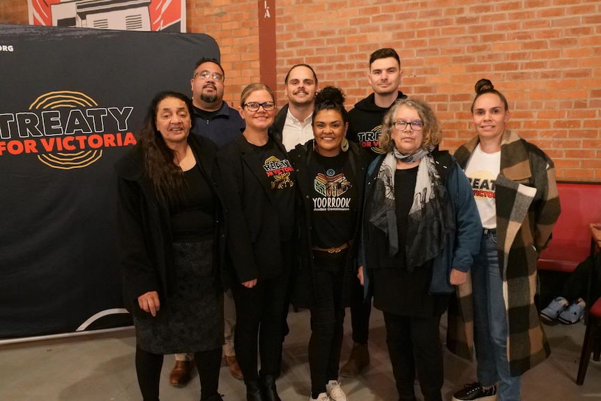 A group of smiling Aboriginal men and women stand together beside a poster reading 'Treaty for Victoria'.