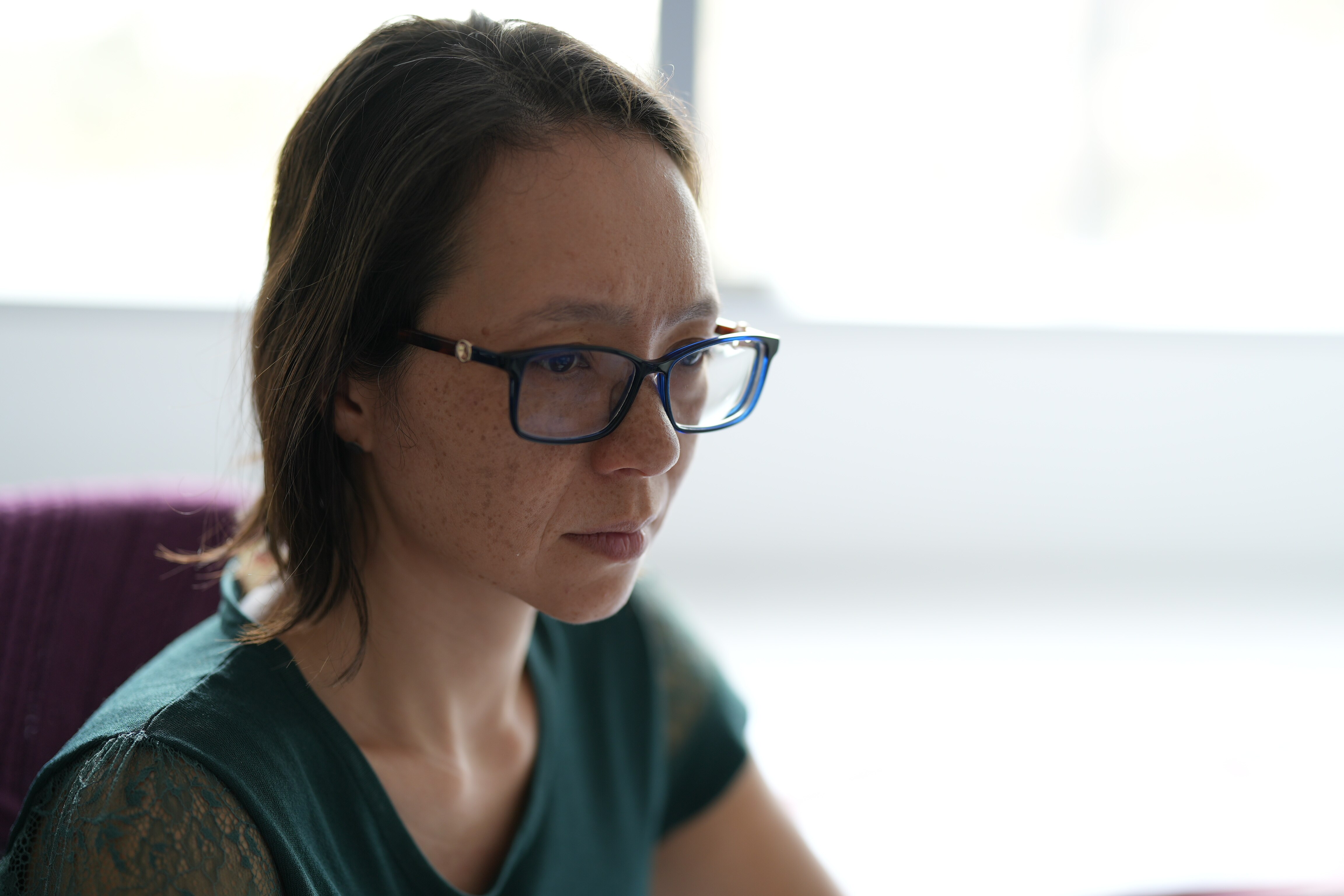 Mulher com corte de cabelo bob, magra e morena, vestindo blusa verde, sentada a uma escrivaninha (fora da vista), óculos e expressão concentrada.