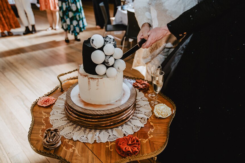 Two people are seen holding one knife and plunging it into a black and white wedding cake on a small table.