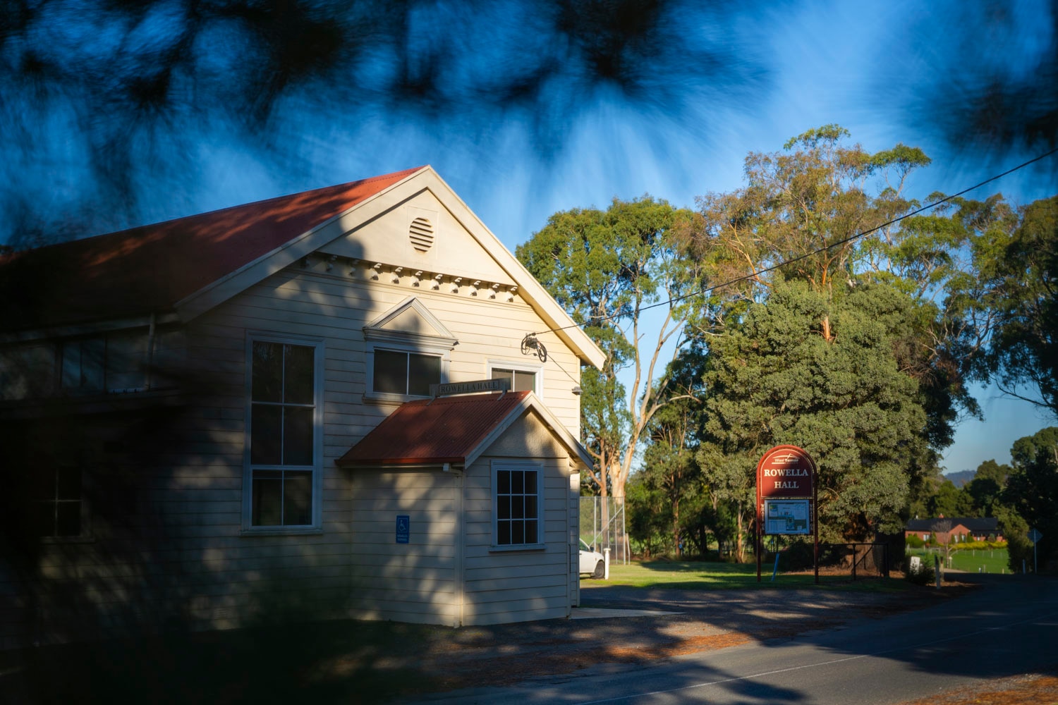 a white one hundred year old wooden clad church-like building sits amongst some greenery with a red sign