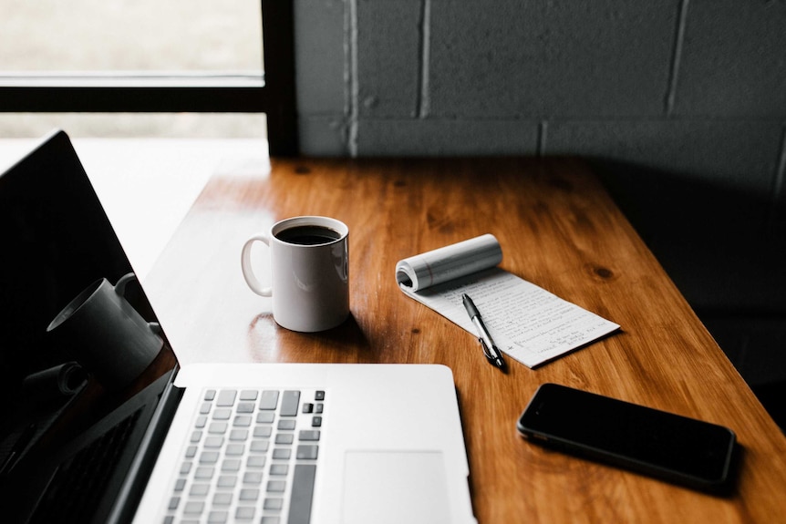 A laptop, coffee, phone and pen and pad on a wooden desk