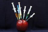 An apple with multiple syringes sticking out of it.