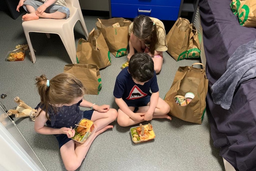 Three children sit on the floor eating food from plastic containers.