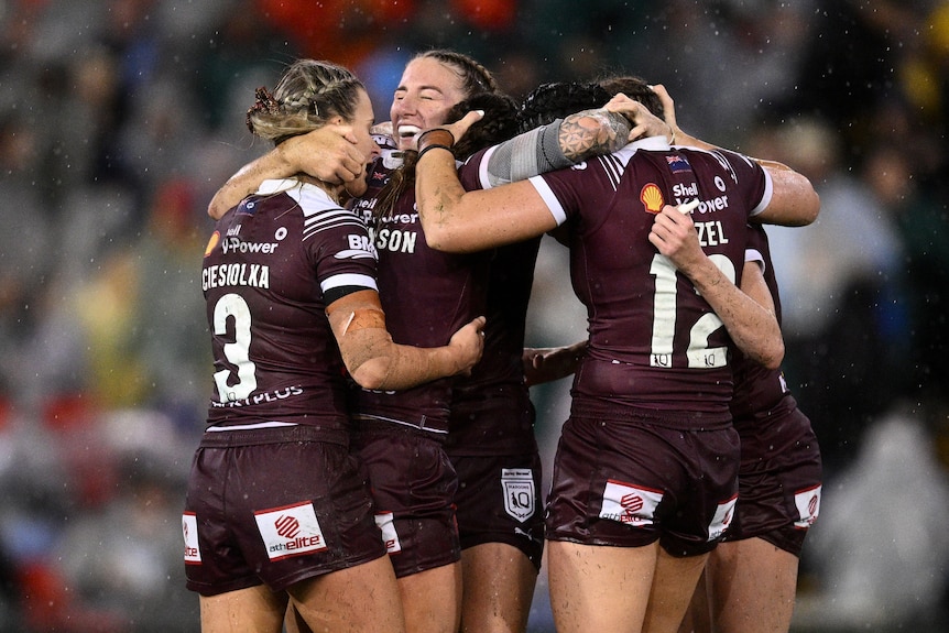 Six members of the women's QLD Maroons embrace in a group hug, in pouring rain, after winning State of Origin Game II