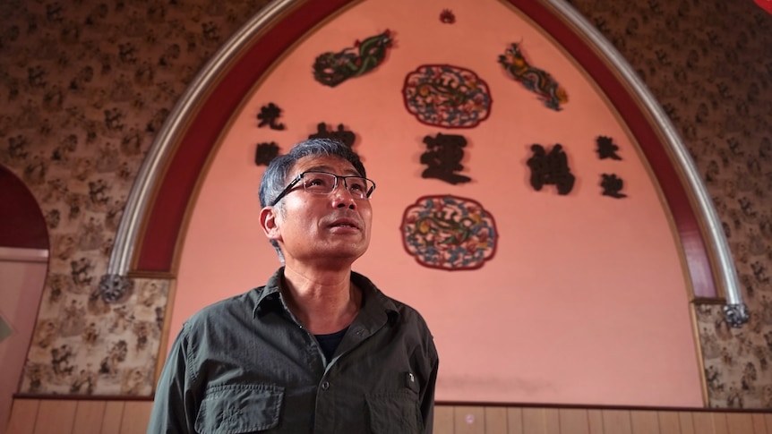 A man looking up at the ceiling, behind him is a wall decorated with Chinese characters and images.