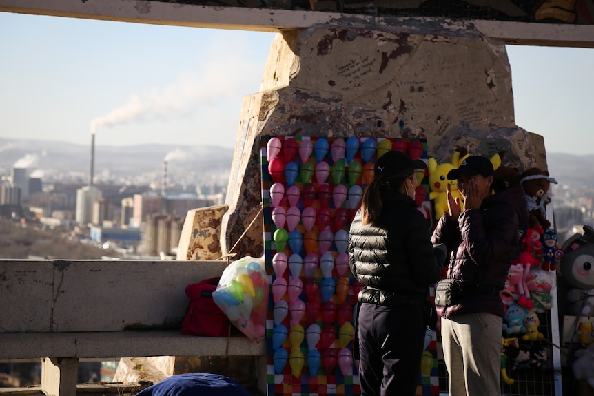 Two people stand in front of a stand with colourful balloons and toys, in front of a lookout overseeing a city