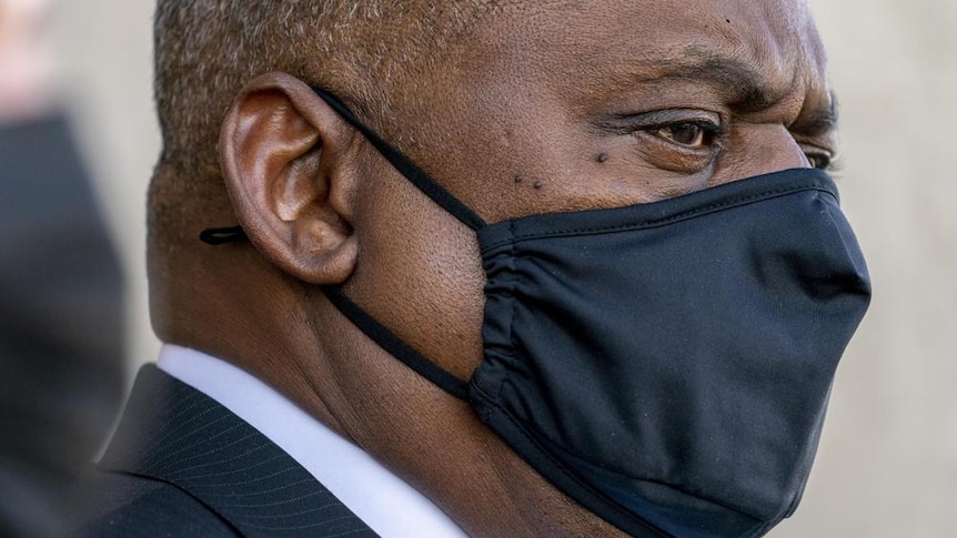 A man wearing a black face mask looks out with a stern facial expression 