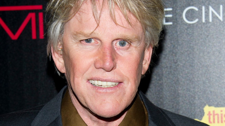 Gary Busey smiles for a photo in front of a movie premiere backdrop. He is wearing a dark suit and blue tie. 