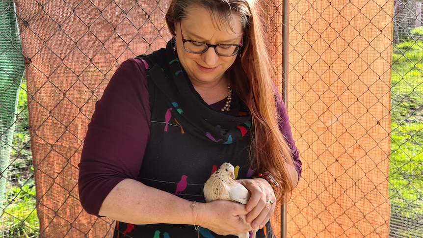 Angela is a woman with long hair, holding a pale-coloured quail in her hands. Angela's head is bowed looking at the bird.