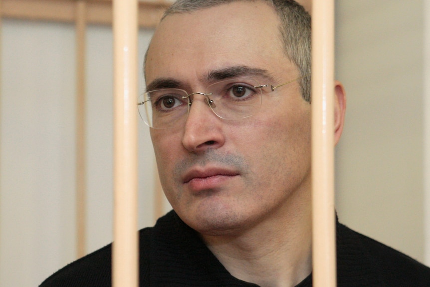 A man with white hair and wearing glasses stares in the distance while behind bars.