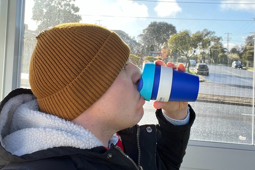 Sabastian wears a winter coat at beanie, drinking from a keep cup on the bus with the view of the sunny street outside.