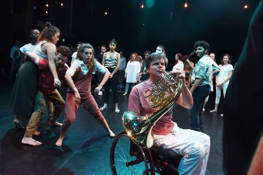 Group of people on stage, man in wheelchair playing French Horn in foreground, cluster of dancers to left. Audience behind.
